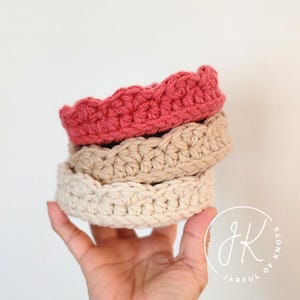 Puede incluir: Una pila de tres diademas de ganchillo hechas a mano en tonos coral, beige y crema. Las diademas están hechas con un punto texturizado y tienen un borde festoneado. Las diademas están apiladas una encima de otra.