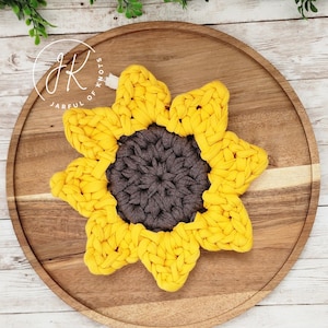 May include: A handmade yellow and grey sunflower coaster made of thick, woven fabric. The coaster is in the shape of a sunflower, with yellow petals surrounding a grey centre. The coaster is placed on a wooden tray.