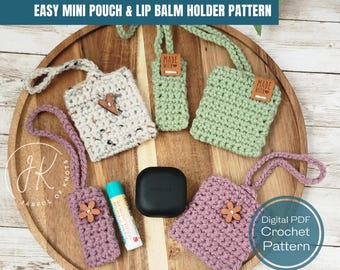 May include: A digital PDF crochet pattern for making easy mini pouches and lip balm holders. The image shows four crocheted pouches in different colors and styles, including a white and gray pouch with a heart, a green pouch with a "Made With" tag, a pink pouch with a flower, and a pink pouch with a flower. The pouches are displayed on a wooden tray with a lip balm tube and a small black container.