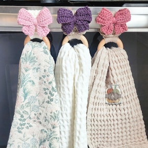 May include: Three decorative kitchen towel holders with wooden rings and crocheted bow accents in pink and purple. The towels are in cream, beige, and a floral pattern. The towels are hanging on a metal bar.