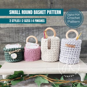 May include: Four crocheted round baskets in different colors, including green, pink, beige, and gray. Each basket has a wooden handle and a small tag that says "Hand Made". The baskets are displayed on a white wooden shelf with green foliage in the foreground.