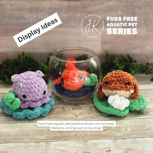 May include: A display of three crocheted aquatic pet toys. A purple hippo sits on a teal and green lily pad. An orange fish rests in a glass bowl. A brown frog sits on a green lily pad. The text reads "Display Ideas" and "Fuss Free Aquatic Pet Series."