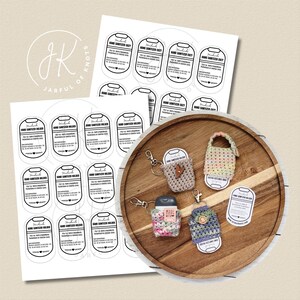 May include: A wooden tray displays handmade hand sanitizer holders and labels. The holders are crocheted in various colors and patterns, with keychains attached. Printed labels with text are also visible, along with the logo "Jarful of Knots".