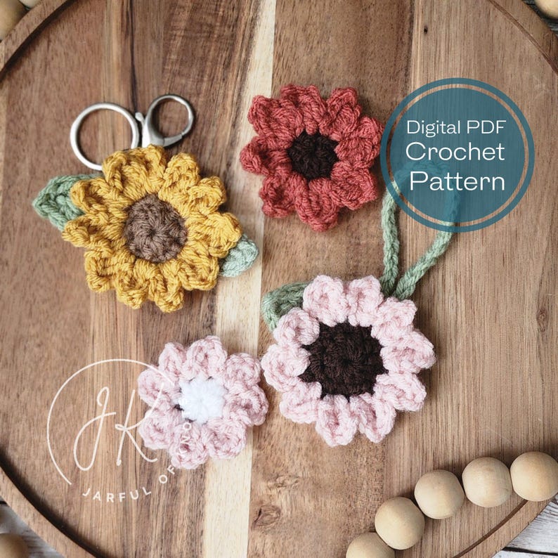 May include: A digital PDF crochet pattern for making three different flower keychains. The keychains are crocheted in yellow, pink, and red with brown centers. The pattern includes instructions for making the flowers and leaves, as well as a keychain loop.