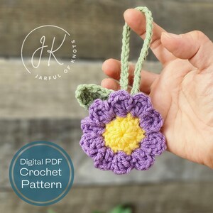 May include: A crocheted purple flower with a yellow center and green leaves. The flower is attached to a green string, which is held by a hand. The image includes the text "Digital PDF Crochet Pattern".