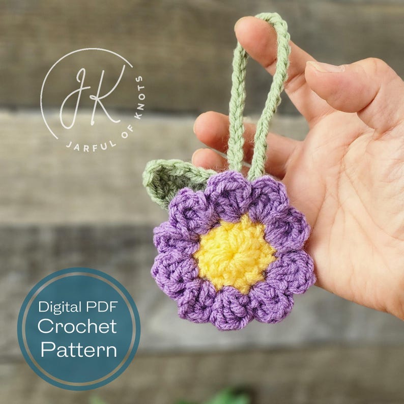 May include: A crocheted purple flower with a yellow center and green leaves. The flower is attached to a green string, which is held by a hand. The image includes the text "Digital PDF Crochet Pattern".