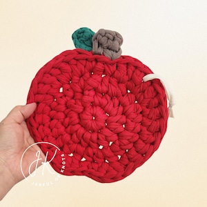 Puede incluir: Un salvamanteles hecho a mano de ganchillo rojo con forma de manzana, con detalles de tallos verdes y grises. El salvamanteles es redondo y tiene un lazo de cuerda blanco. El producto está hecho de hilo grueso y texturizado.