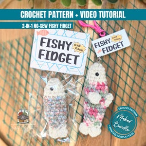 May include: A crochet pattern and video tutorial for a 2-in-1 no-sew fishy fidget. The set includes a small crocheted fish in a clear plastic bag with a tag labeled Fishy Fidget, and a larger crocheted fish attached to a pink plastic clip.