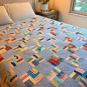 May include: A patchwork quilt featuring a repeating pattern of light blue fabric squares interspersed with multi-colored, patterned fabric blocks. The quilt is stitched with a fine grid texture and covers a bed, displaying various textile prints.