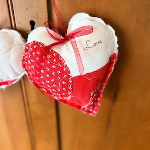 May include: A handmade heart-shaped ornament with a patchwork design. The ornament features red and white fabric, with a red ribbon bow and a tag that says "Love". The ornament is hanging from a red ribbon.