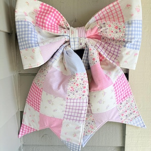 May include: A large, decorative bow made of patchwork fabric. The bow features squares of pink gingham, floral prints, and solid pink, purple, and blue fabrics. The bow is designed to be hung and adds a touch of charm to any space.