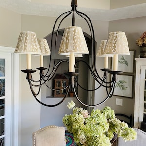 May include: A bronze-colored chandelier with five cream-colored lampshades featuring a spotted pattern. The chandelier hangs above a dark wooden table with a vase of green and pink flowers. The room has a neutral color palette.
