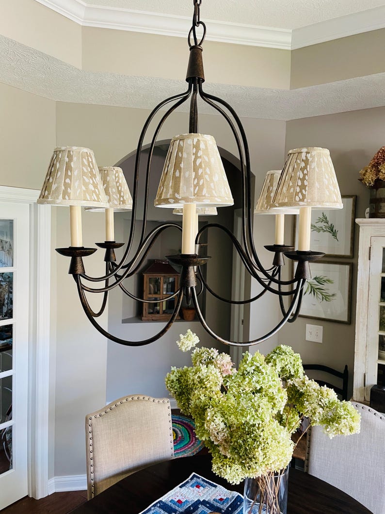 May include: A bronze-colored chandelier with five cream-colored lampshades featuring a spotted pattern. The chandelier hangs above a dark wooden table with a vase of green and pink flowers. The room has a neutral color palette.