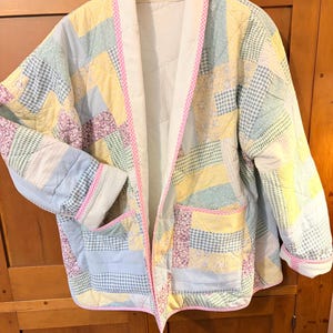 May include: A quilted jacket featuring a patchwork design of pastel yellow, light blue, and lavender fabric strips with floral and gingham patterns. The jacket has a white collar, pink and white checkered trim, and two front pockets.