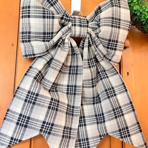 May include: A large decorative bow with a plaid pattern in black, white, and gray. The bow is hanging on a white hook against a wooden door. The bow's fabric is folded and tied in the center.