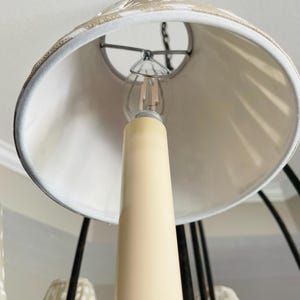 May include: Close-up of a chandelier with a cream-colored candle-shaped light bulb holder. The lampshade is white with a decorative trim. The chandelier has a dark metal frame and is hanging from the ceiling.