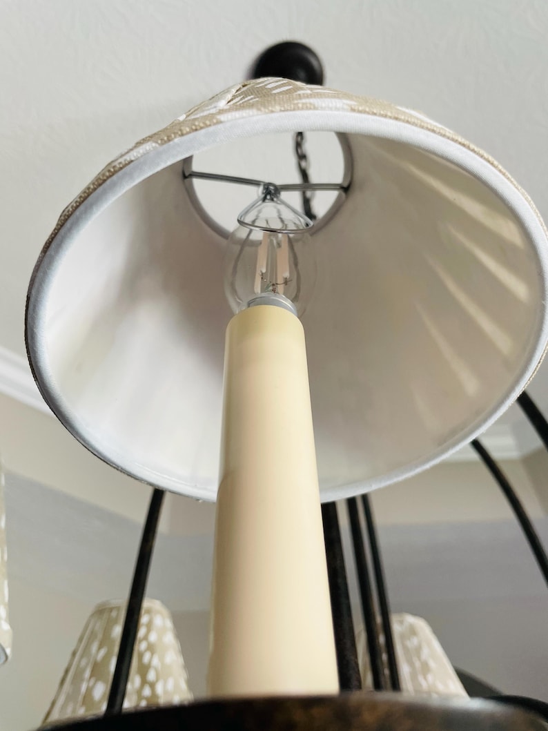 May include: Close-up of a chandelier with a cream-colored candle-shaped light bulb holder. The lampshade is white with a decorative trim. The chandelier has a dark metal frame and is hanging from the ceiling.