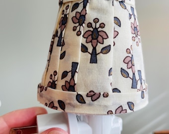 May include: A small, fabric lampshade with a floral pattern in pink, blue, and gold on an off-white background. The lampshade is attached to a white plug-in night light.
