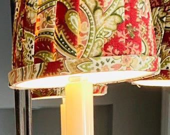 May include: Close-up of a chandelier with pleated lampshades. The lampshades feature a red paisley pattern with green and beige accents. The chandelier has a dark metal frame and candle-shaped lights.