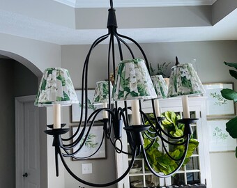 May include: Black wrought iron chandelier with six candle-style lights and floral patterned shades. The shades feature a white background with green leaves and flowers. The chandelier hangs from a black chain.