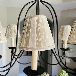 May include: A bronze chandelier with multiple arms, each holding a cream-colored candle. The lampshades are pleated and beige with a white spotted pattern. The chandelier is in a room with neutral-colored walls and a white ceiling.