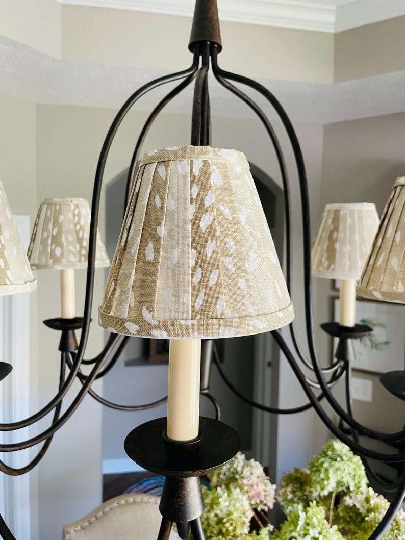 May include: A bronze chandelier with multiple arms, each holding a cream-colored candle. The lampshades are pleated and beige with a white spotted pattern. The chandelier is in a room with neutral-colored walls and a white ceiling.
