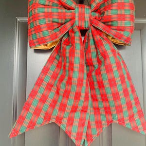 May include: A large, decorative bow in a classic plaid pattern of red, green, and tan. The bow is made of fabric and has a gold-colored lining. It is designed for door or wall decor, perfect for the holiday season.