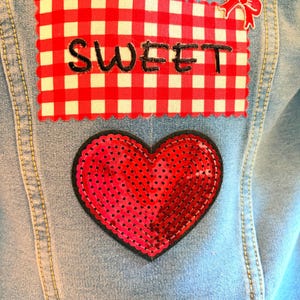 May include: A light blue denim jacket featuring a rectangular patch with a red and white gingham pattern, the word SWEET embroidered in black, and a small red bow applique. Below is a heart-shaped patch covered in shiny red sequins.