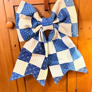 May include: A large, decorative bow made of blue and white patchwork fabric. The bow is suspended by a brown leather strap and a metal ring. The fabric features various patterns, including polka dots and squares. The bow is likely used for home decor.