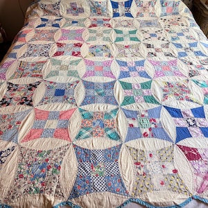 May include: A patchwork quilt featuring a repeating pattern of square blocks in various floral, plaid, and geometric prints. The blocks are separated by cream-colored fabric segments with curved edges, and the entire quilt has a scalloped blue border.
