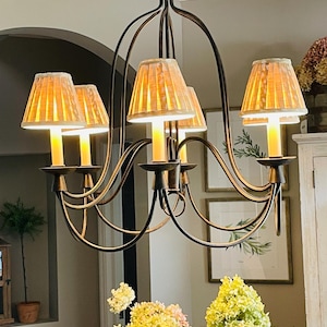 May include: A bronze-colored chandelier with five candle-style lights and pleated lampshades. The fixture has curved arms and a central support. The lampshades are a light beige color. The chandelier is hanging in a room with framed artwork and a floral arrangement.