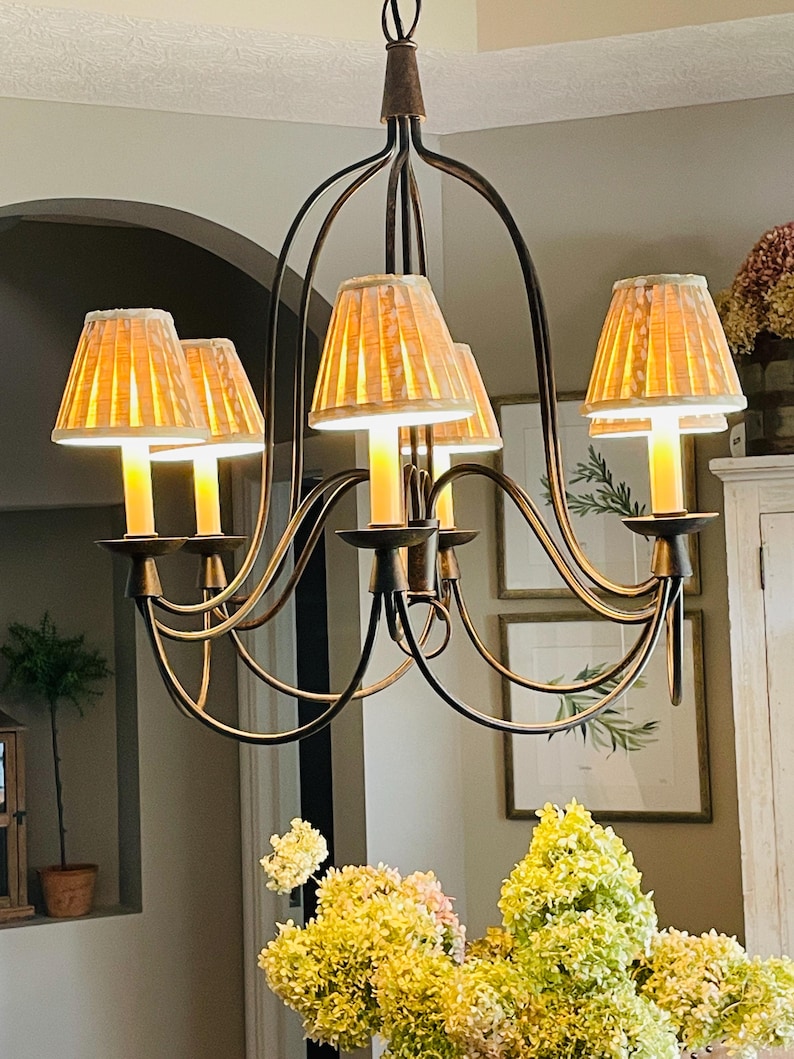 May include: A bronze-colored chandelier with five candle-style lights and pleated lampshades. The fixture has curved arms and a central support. The lampshades are a light beige color. The chandelier is hanging in a room with framed artwork and a floral arrangement.