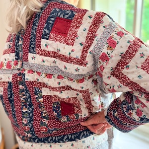 May include: A quilted jacket featuring a patchwork log cabin pattern in shades of deep red, navy blue, and white. The fabric displays various floral and geometric prints, with visible stitching throughout the garment and a solid red trim at the hem.