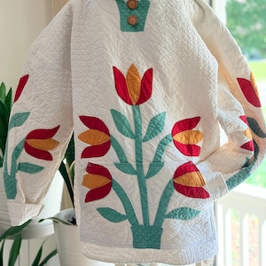 May include: A white quilted hooded pullover featuring a large floral appliqué design with red and orange tulips on green stems. The garment includes a front kangaroo pocket, long sleeves, and two round wooden buttons at the neckline collar.