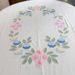 May include: A white tablecloth featuring an oval-shaped floral embroidery pattern. The design includes pink, light blue, and pale yellow fabric flower appliqués with sage green leaves, all outlined with decorative stitching on a plain white fabric.