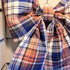 May include: A large decorative bow made of fabric featuring a plaid pattern in shades of blue, orange, white, and black. The bow has two wide loops and two long, hanging tails, all constructed from the same tartan-style textile material.