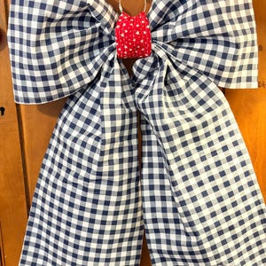 May include: A large decorative bow made of blue and white gingham fabric. The bow is centered with a red fabric piece adorned with white stars. A brown leather strap and metal ring are attached for hanging.