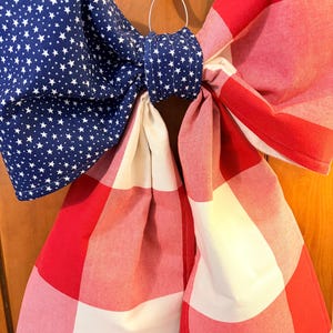May include: A patriotic bow featuring a blue fabric with white stars and red and white checkered fabric. The bow is tied with a blue fabric ring and hangs from a metal ring.