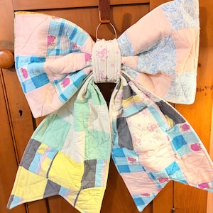 May include: A large, quilted bow in pastel colors, including pink, blue, yellow, and green. The bow is made of patchwork fabric and has a brown leather strap for hanging. The bow is a decorative item.