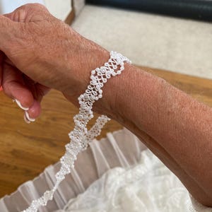 May include: A white beaded bracelet with a delicate, intricate design. The bracelet is made of small, white beads that are strung together in a pattern of loops and knots. The bracelet is shown on a person's wrist.