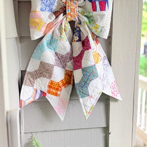 May include: A large decorative bow constructed from quilted fabric patches featuring a variety of colorful floral, plaid, and geometric patterns. The bow is tied in the center with a matching fabric band and has two long, pointed tails hanging down.