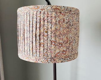 May include: A cylindrical lamp shade featuring a pleated fabric design with a repeating floral pattern in shades of peach, pink, and white. The shade is mounted on a dark metal stand and has a structured, uniform shape with visible vertical folds.