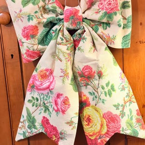 May include: A large decorative bow with a floral pattern featuring pink, red, and yellow roses on a cream background. The bow is made of fabric and has a loop for hanging. Green leaves and stems are also visible.