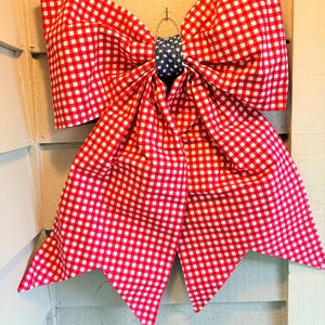 May include: A large decorative bow made of red and white gingham fabric. The center of the bow features a blue fabric with white stars. The bow is hanging on a light-colored wooden surface.