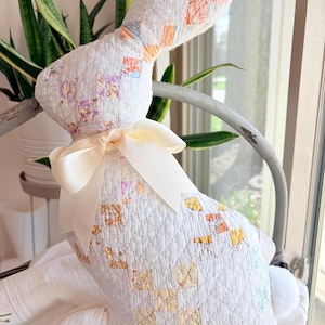May include: A handmade white fabric bunny with a cream-colored bow around its neck. The bunny is made from a patchwork quilt with pastel-colored squares. It is sitting in a metal basket with a white cloth.