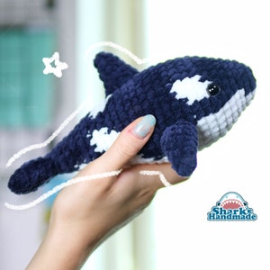 Op de afbeelding: Een gehaakt pluche speelgoed in de vorm van een orka. De orka is blauw met witte vlekken en heeft een witte buik. Het speelgoed wordt in een hand gehouden.