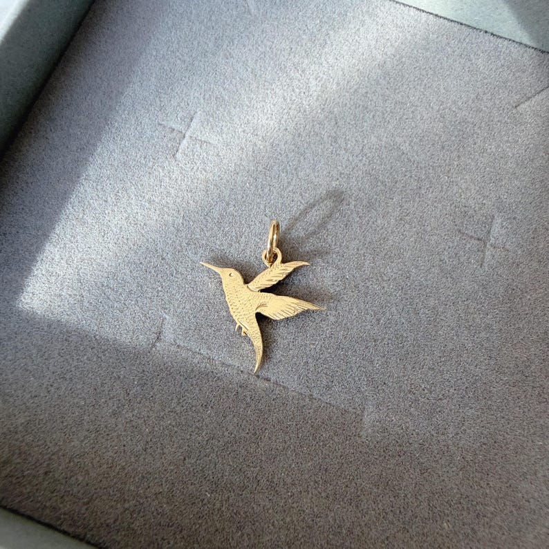 May include: A gold-colored metal pendant shaped like a hummingbird in flight, featuring detailed etched lines on the wings and body to represent feathers, with a small circular jump ring attached at the top for hanging on a chain or necklace.