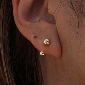 以下が含まれることがあります： 耳たぶに装着された2つの光沢のあるゴールドトーンのボール型スタッドピアス。各ピアスは、反射するメタリック仕上げの滑らかな球体デザインが特徴です。スタッドは上下に配置されており、シンプルで洗練された外観を作り出しています。