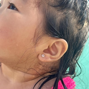 以下が含まれることがあります： 小さな丸い輝くスタッドピアスをつけた子供の耳のクローズアップ。ピアスは、透明なクリスタルに囲まれた中央のピンク色の石が特徴です。子供の肌は白く、黒い髪は濡れています。