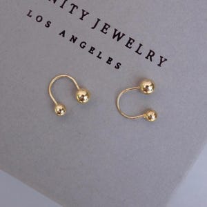 May include: A pair of gold-toned earrings featuring a curved U-shaped wire design with a small polished sphere at each end. The earrings are positioned above the text "UNITY JEWELRY LOS ANGELES" printed in black on a light gray surface.